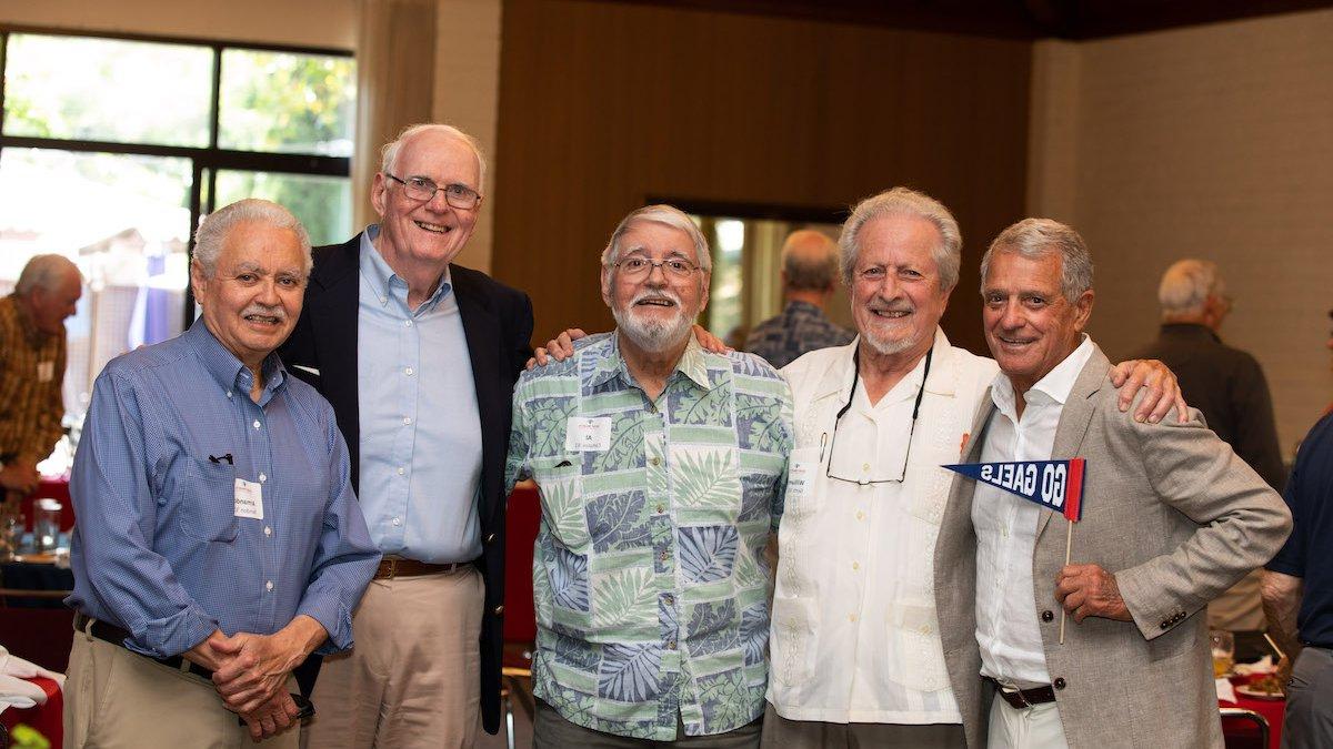 Five alumni smile at the camera, one holding a 盖尔人去 pennant